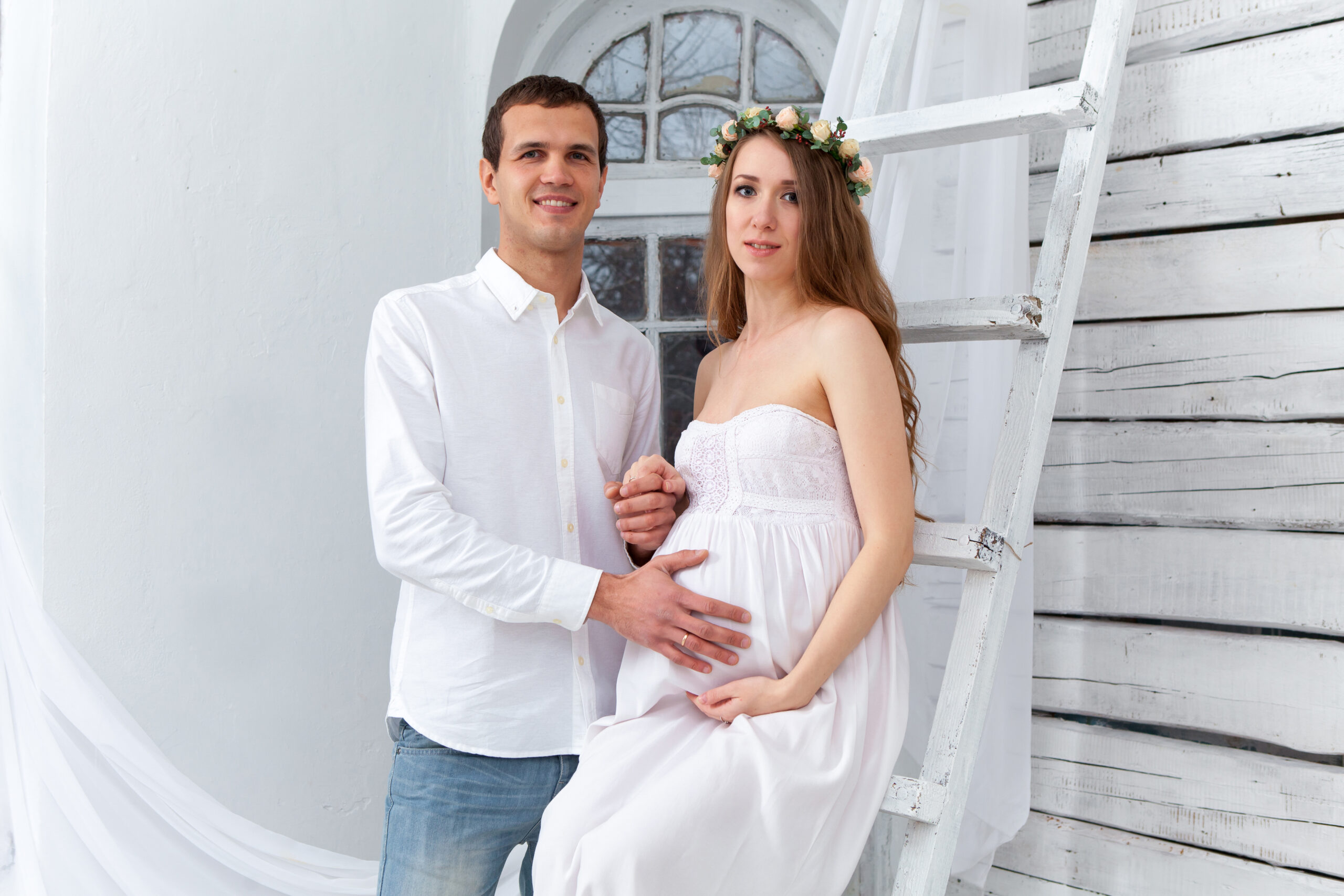 Cheerful young couple dressed in white standing against white home. Husband hugging his pregnant wife